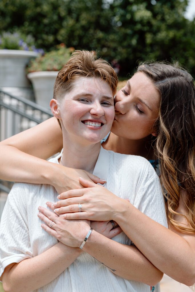 women hugs and kisses woman from behind at queer engagement at Tulsa Botanical Garden