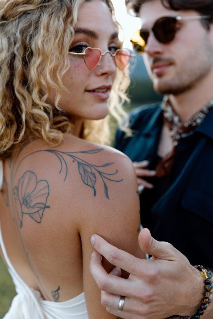 groom holds bride as she looks over shoulder with red heart gold sunglasses with poppy tattoo 
