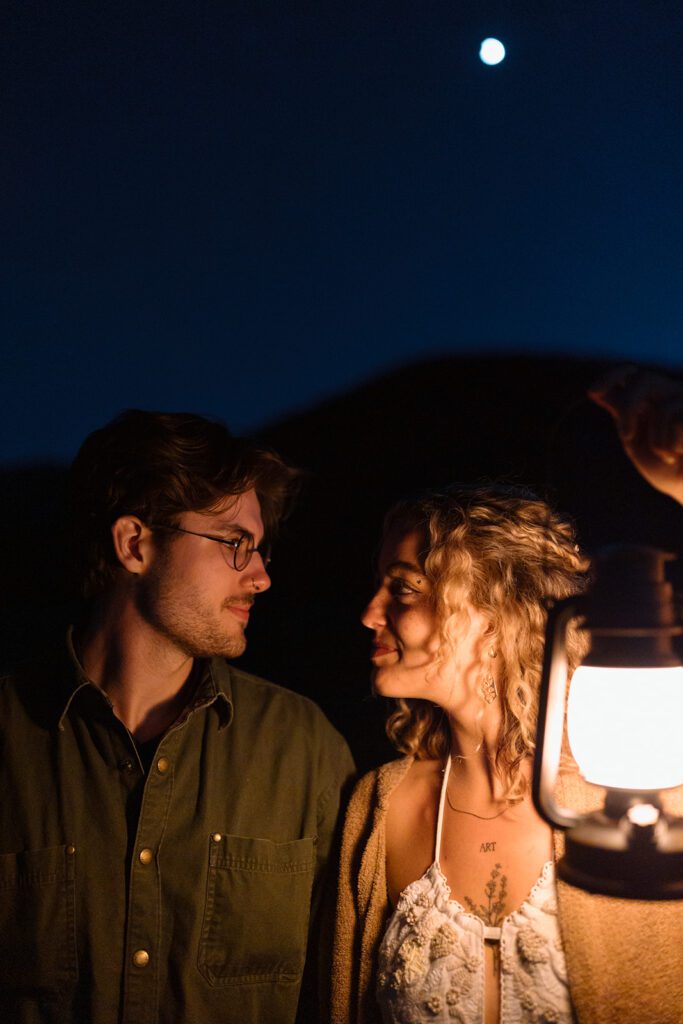 bride looks at groom holding lantern at night during camping elopement in Colorado