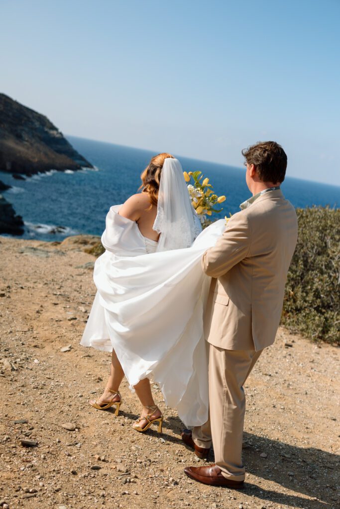 groom carries brides train as the walk along rocky coast in greece 