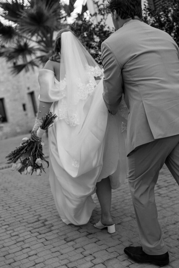 groom carries brides train on cobblestone street in black and white 