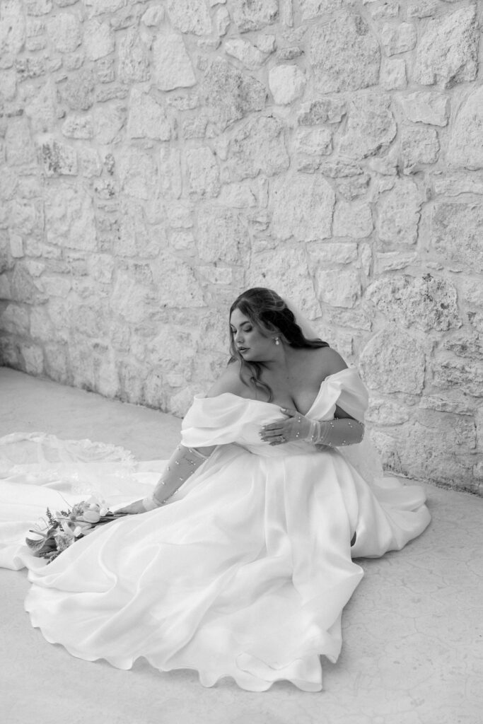 plus size bride sits on floor for portrait with stone wall and bridal bouquet in black and white 