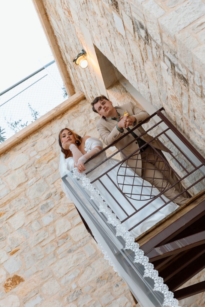 bride and groom look down from metal porch on stone building 