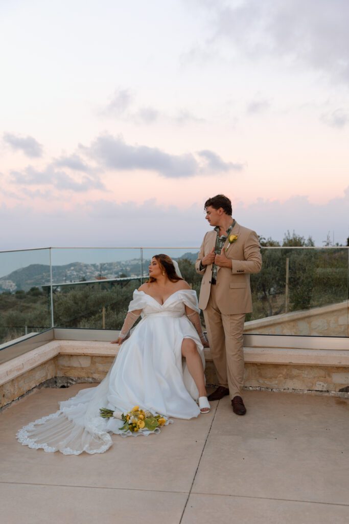 bride sits and groom stands as they look out to sea from patio in Greece